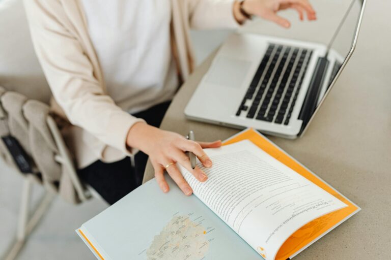 A person sits at a desk reading an open book beside a laptop, focusing on study or work.