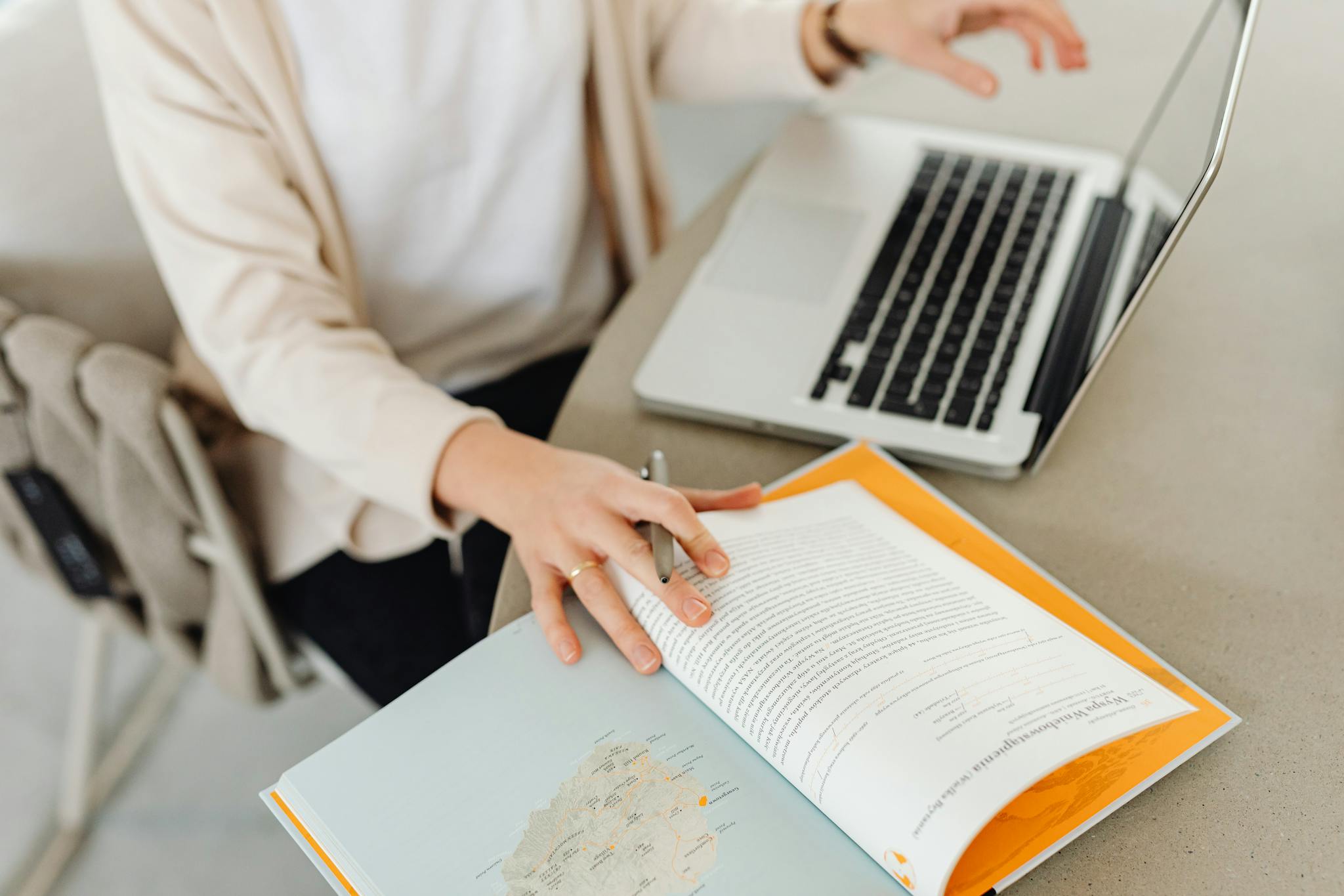 A person sits at a desk reading an open book beside a laptop, focusing on study or work.