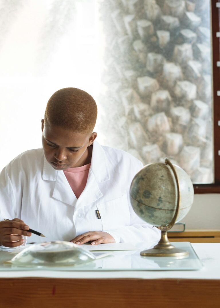 Concentrated astrophysicist in white coat studying constellation materials while sitting at table opposite magnifier and globe in light scientific office